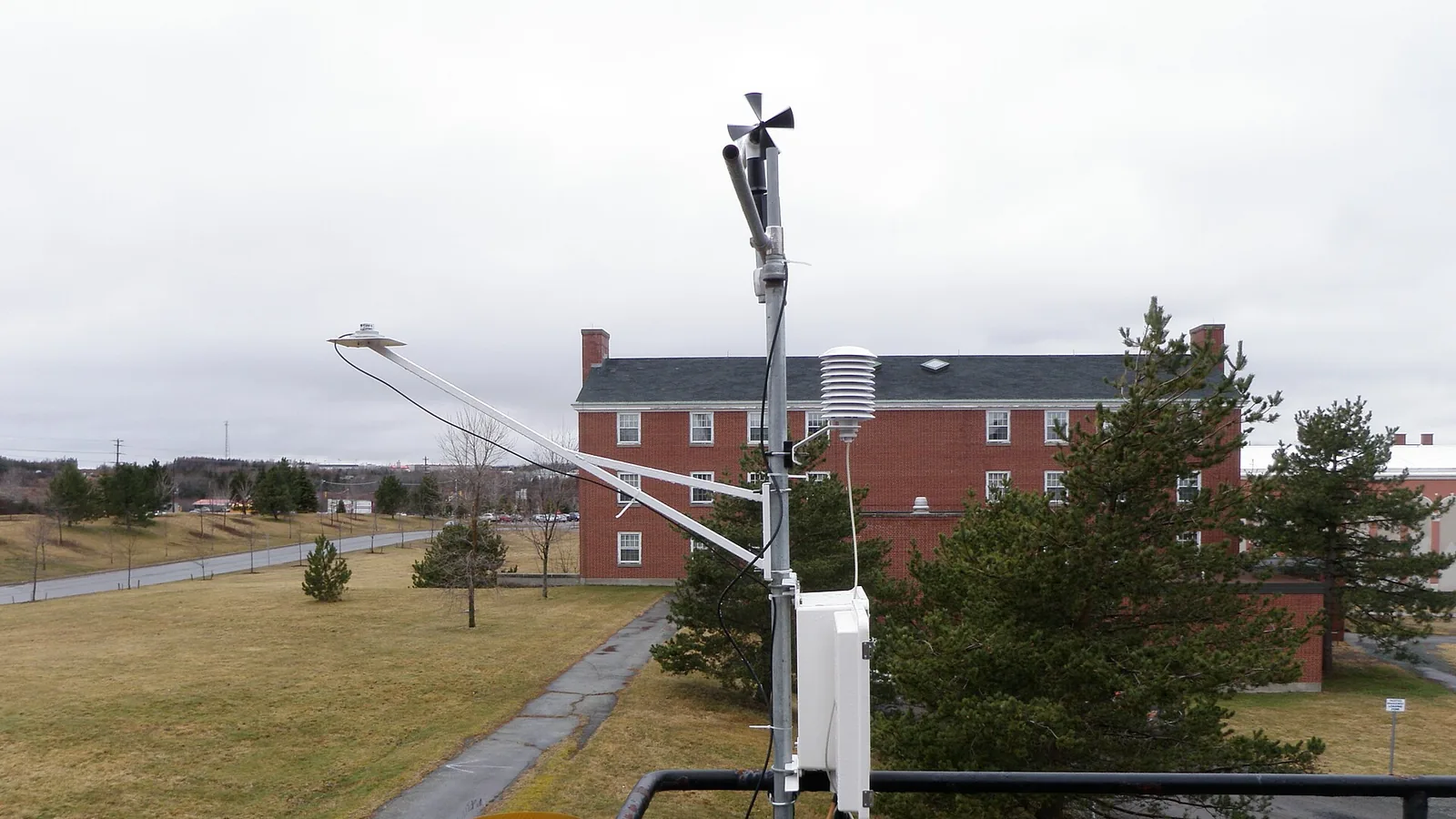 A real outdoor weather station with wind and precipitation instruments beside trees and buildings
